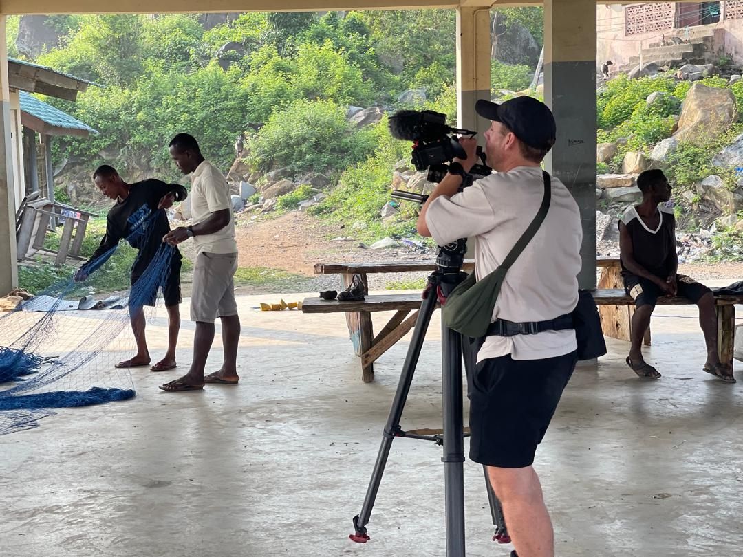 Crew filming fishermen mending nets under a community shelter in Dixcove